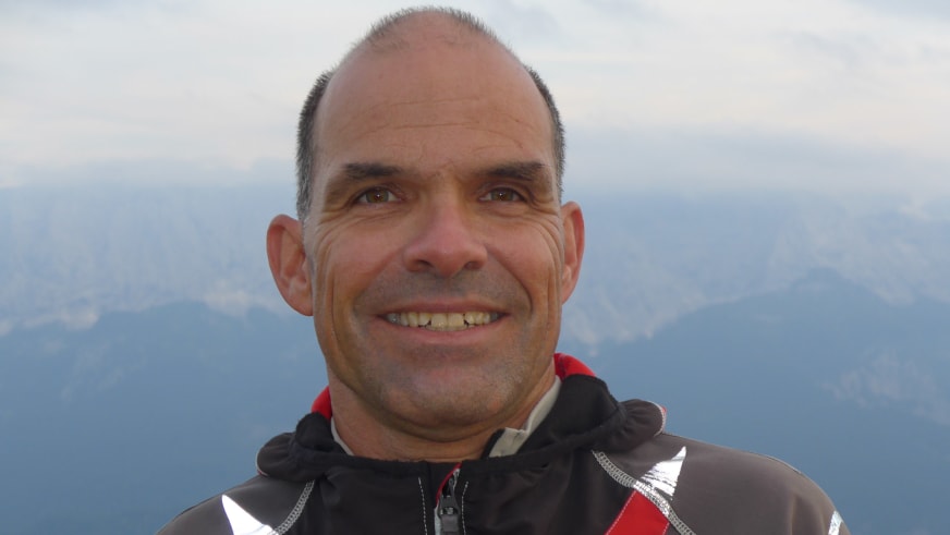 Bergwanderführer Andreas Schneider lächelt in schwarzer Softshell-Jacke vor Berglandschaft mit mehreren Bergketten im Dunst.