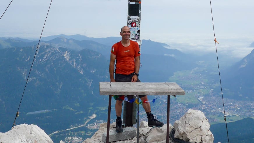Bergwanderführer Andreas Schneider steht hinter hölzernem Tisch auf Felsgipfel mit Gipfelkreuz und Panoramablick über Bergtal.