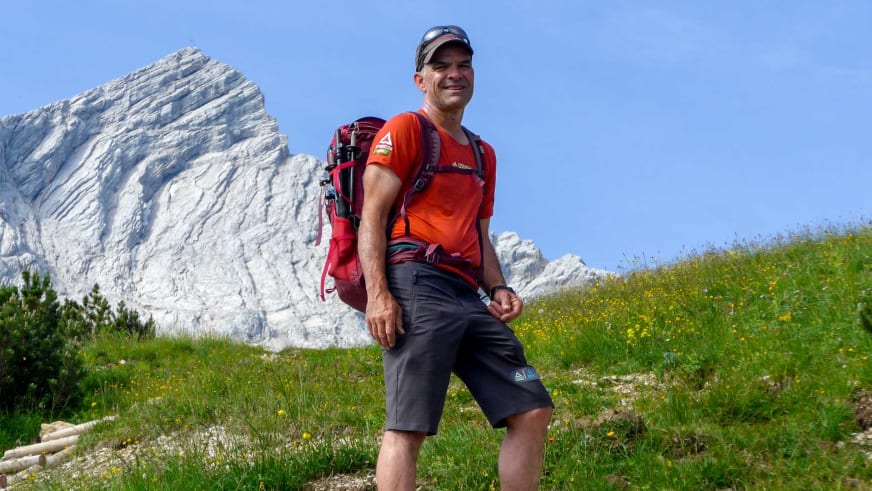 Bergwanderführer Andreas Schneider steht in oranger Funktionskleidung auf einer grünen Wiese mit gelben Blumen vor Kalksteinberg.