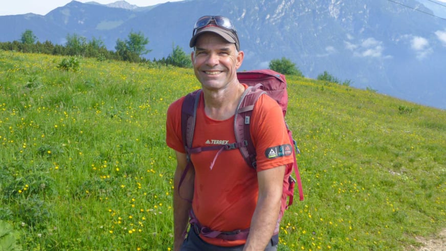 Bergwanderführer Andreas Schneider trägt rotes Terrex-Shirt und Wanderrucksack vor grüner Almwiese und bewaldeten Hängen.