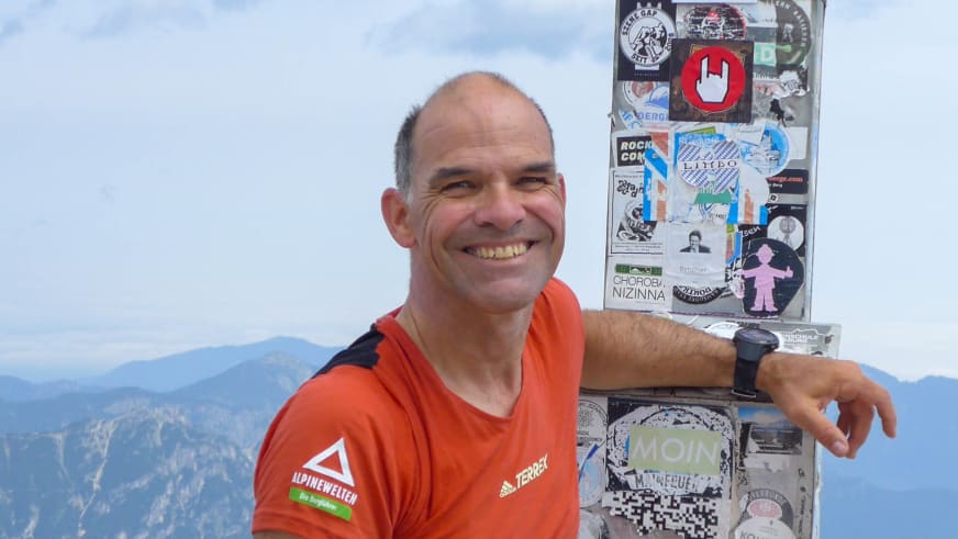 Bergwanderführer Andreas Schneider in orangem T-Shirt lehnt entspannt an einer Metallbox vor alpiner Berglandschaft.
