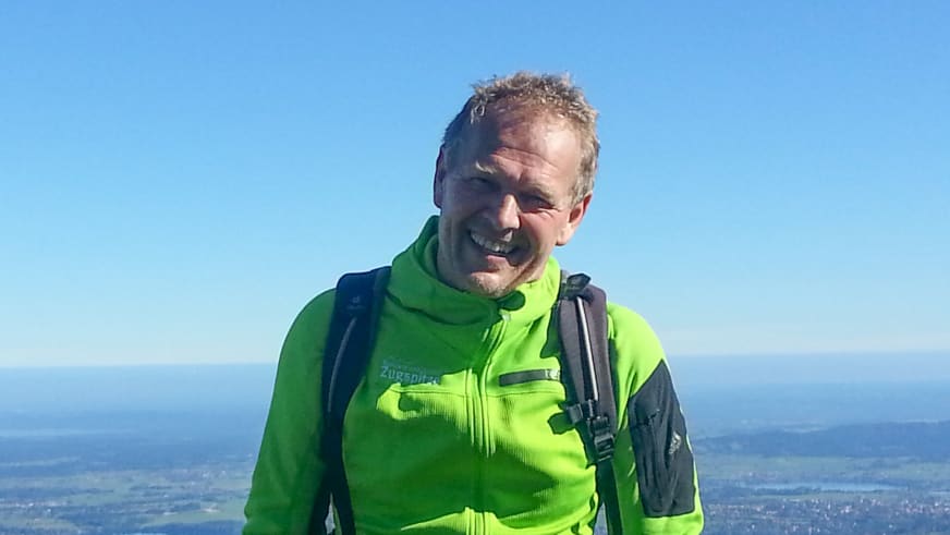 Bergwanderführer Martin Werner trägt leuchtend grüne Zugspitze-Softshell-Jacke mit schwarzem Rucksack vor blauem Himmel.