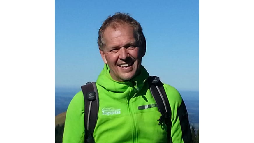 Bergwanderführer Martin Werner lächelt in grüner Bergsportjacke mit Zugspitze-Logo vor blauem Himmel und warmen Berghängen.