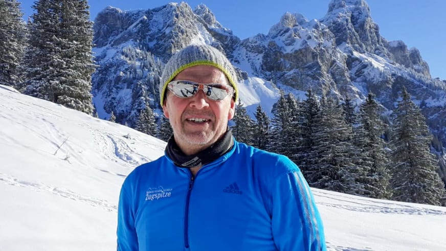 Martin Werner als Bergwanderführer in blauer Zugspitze-Jacke mit Sonnenbrille vor verschneiter Bergkette im Winter.