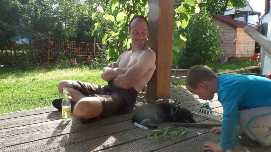 Auf einer Holzterrasse im Garten entspannen sich zwei Personen mit Klettertrainer Volker Wild und einem schwarzen Hund.