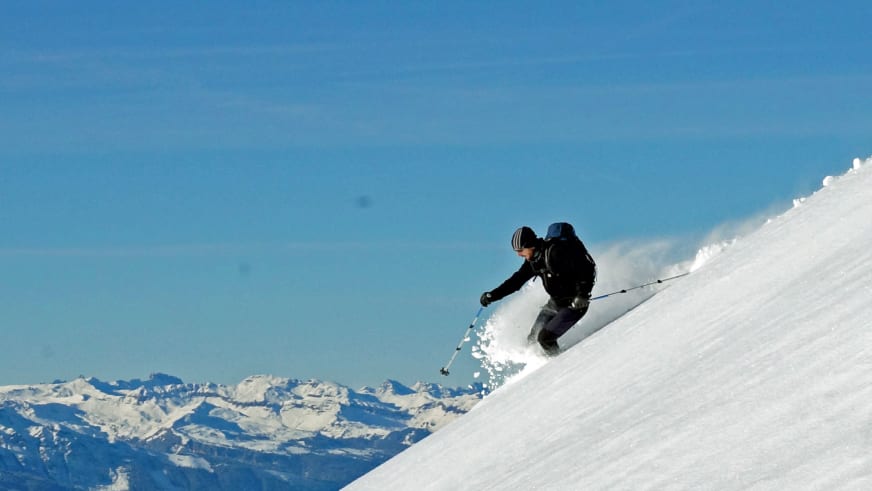 Bergführer Peter Klug fährt auf Skiern einen steilen Schneehang hinab und wirbelt dabei Pulverschnee auf.