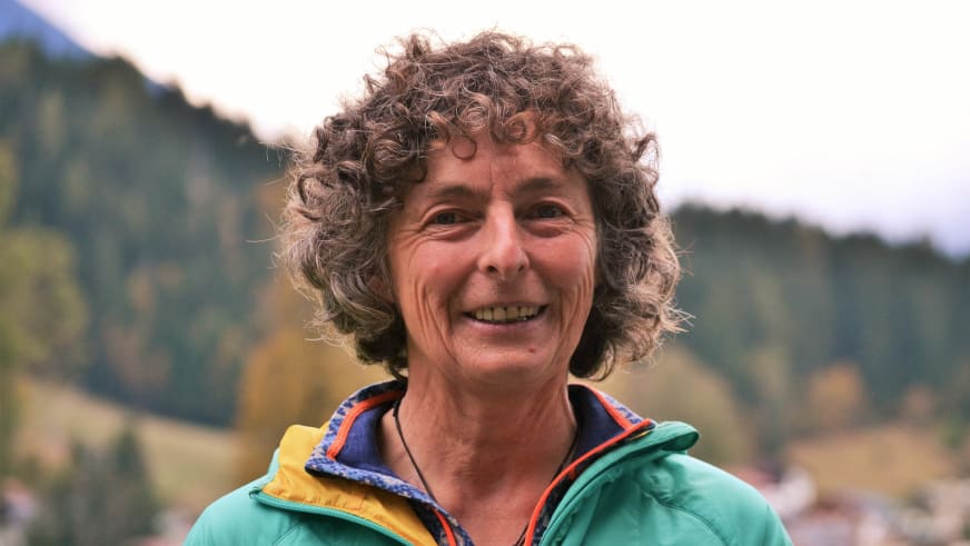 Freundliches Portrait von Bergwanderführerin Irene Aschauer mit lockigem Haar und türkiser Outdoorjacke vor Berglandschaft.