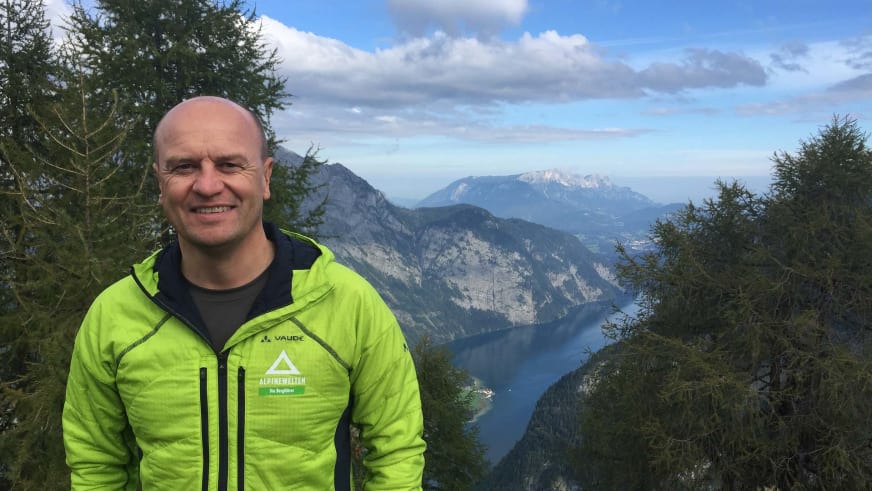 In gelber Vaude-Outdoorjacke lächelt Bergwanderführer Peter Kohl vor einem Bergpanorama mit langgestrecktem See im Tal.