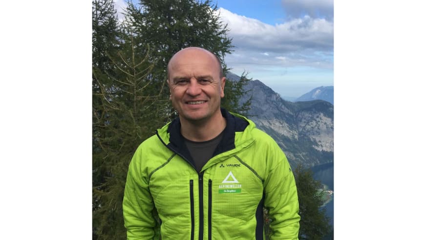 Bergwanderführer Peter Kohl lächelt in grüner Vaude-Jacke vor einer Berglandschaft mit bewaldeten Hügeln und Felswänden.