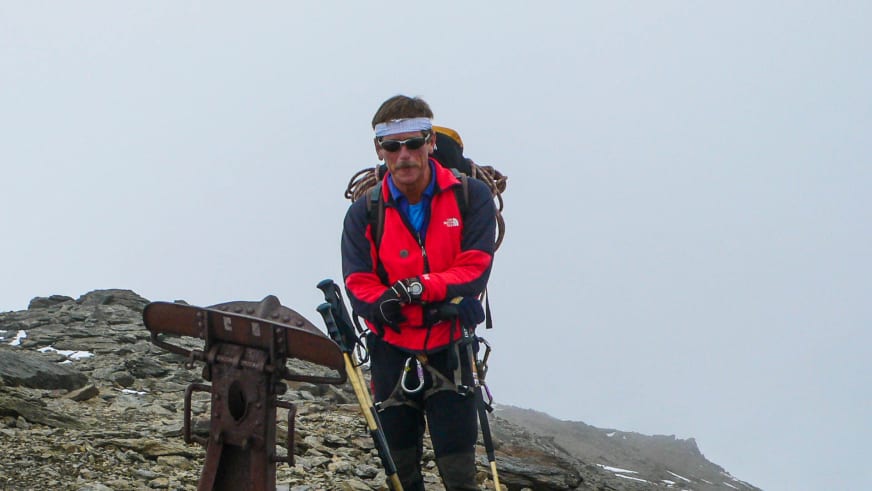 Bergführer Georg Gruber steht mit Wanderstöcken und Rucksack neben einem Metallkreuz auf einem felsigen Gipfel.