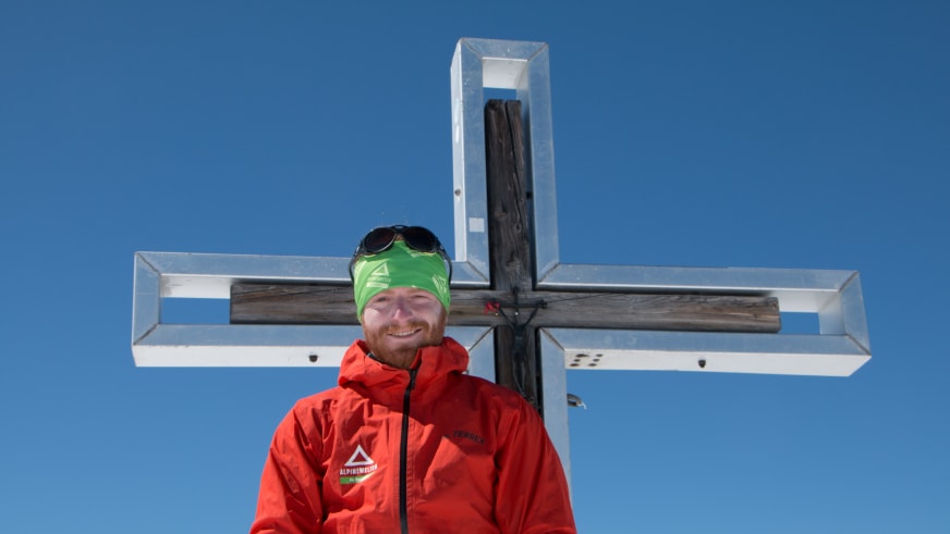 Bergwanderführer Josef Aschauer steht lächelnd vor weißem Gipfelkreuz mit roter Alpinwelten-Jacke und grünem Stirnband bei klarem Himmel.