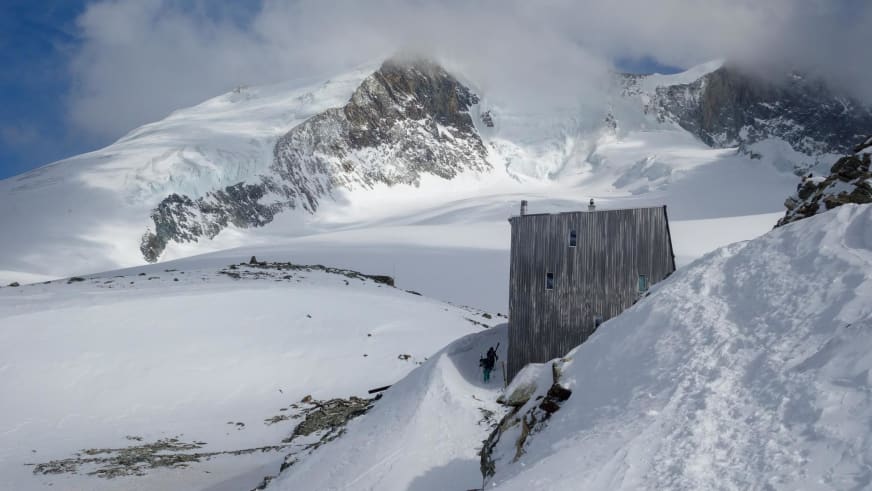 Ein Bergsteiger steht vor der Cabane Tracuit, die auf einem schneebedeckten Grat in der Schweiz liegt.