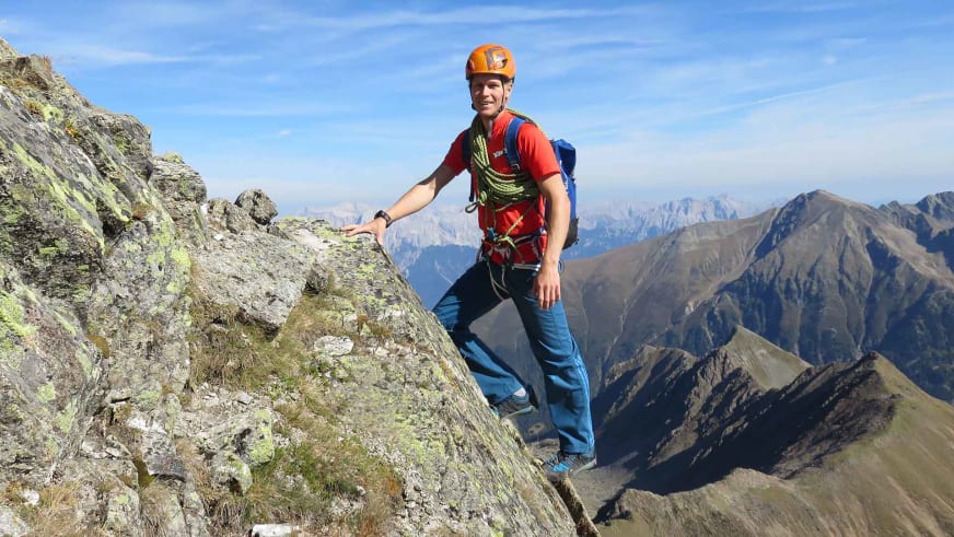 Bergführer Martin Schidlowski klettert mit orangem Helm und gelbem Kletterseil an steiler Felswand vor Bergpanorama.