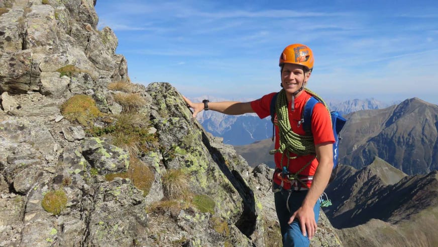 Bergführer Martin Schidlowski steht mit orangem Kletterhelm und grünem Seil lächelnd auf einem Felsgrat in den Bergen.