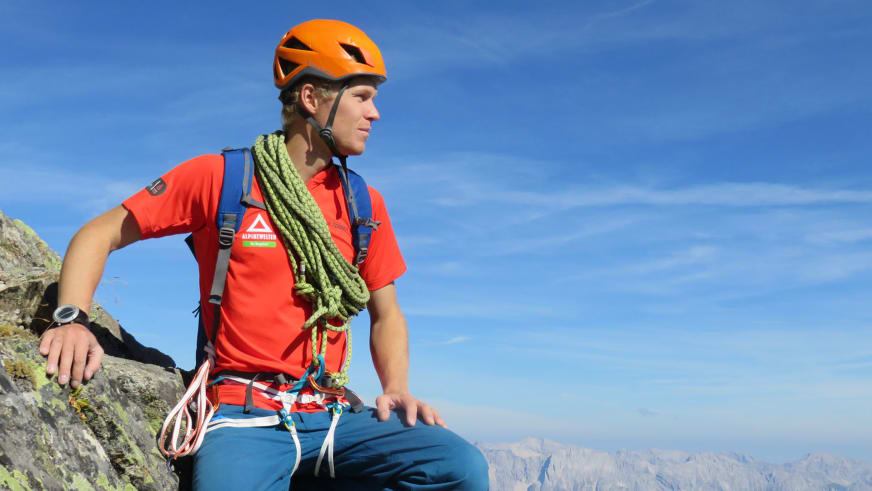 Bergführer Martin Schidlowski sitzt auf einem Felsvorsprung mit rotem Alpine Welten Shirt, orangefarbenem Kletterhelm und Kletterausrüstung.