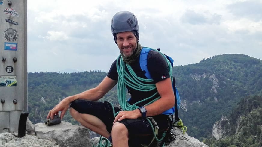 Michael Aigner sitzt entspannt auf einem Felsvorsprung mit blauem Helm, türkisem Kletterseil und Bergführer-Ausrüstung.