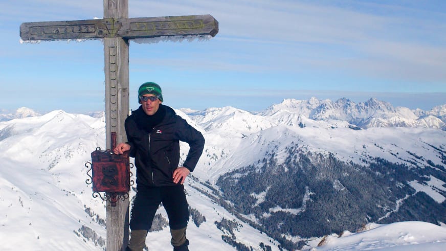Bergführer Michael Aigner steht mit grüner Mütze und Sonnenbrille neben hölzernem Gipfelkreuz auf verschneitem Berggipfel.