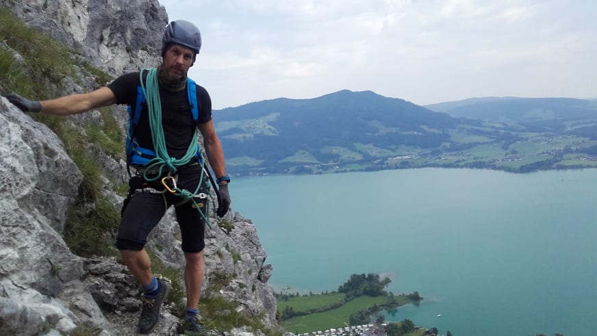 Bergführer Michael Aigner steht mit kompletter Kletterausrüstung auf einem Felsvorsprung oberhalb türkisgrüner Seen in bewaldeter Landschaft.