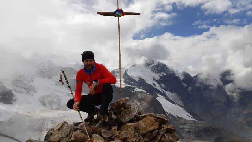 Am Kaunergrat im Pitztal hockt Bergführer Michael Aigner mit Pickel neben Steinmännchen und Gipfelkreuz bei Wolkenstimmung.