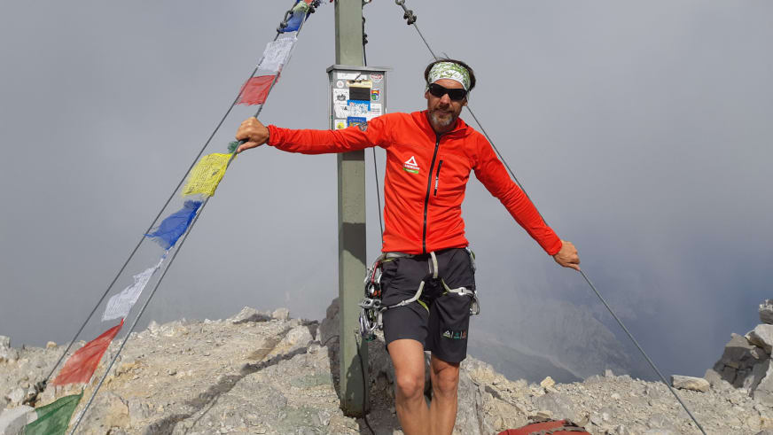 Bergführer Michael Aigner posiert mit ausgebreiteten Armen vor einem Gipfelkreuz mit bunten Gebetsfahnen bei der Watzmann Überschreitung.