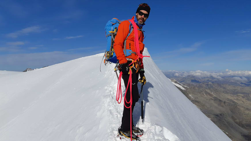 Bergführer Michael Aigner steht mit Steigeisen und orangener Jacke auf schmalem, schneebedecktem Gipfelgrat beim Aufstieg zum Piz Bernina.