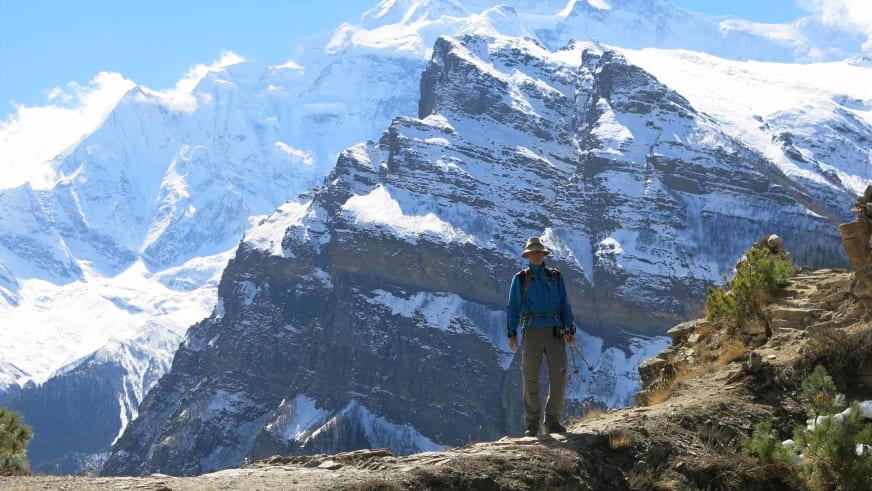 Bergwanderführer Werner Neubauer steht in blauer Jacke und braunem Hut auf steinigem Felsplateau vor schneebedeckten Gipfeln.