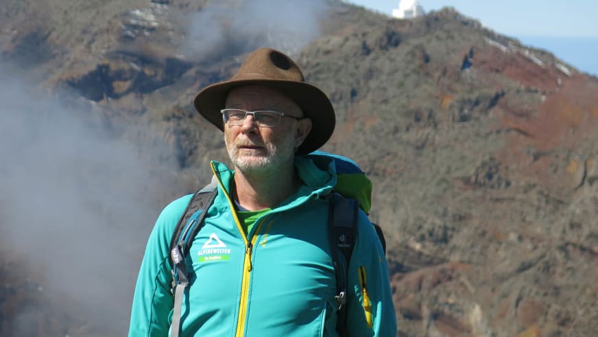 Bergwanderführer Werner Neubauer mit grauem Vollbart und türkiser Softshelljacke vor steilen Felshängen und Nebelschleiern.