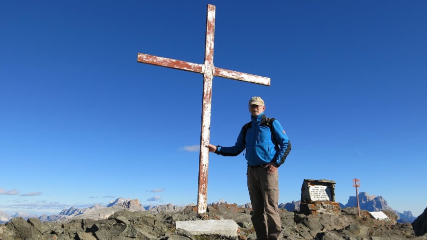Am weißen Gipfelkreuz steht Bergwanderführer Werner Neubauer in blauer Jacke mit Baseballkappe und Sonnenbrille bei klarem Himmel.