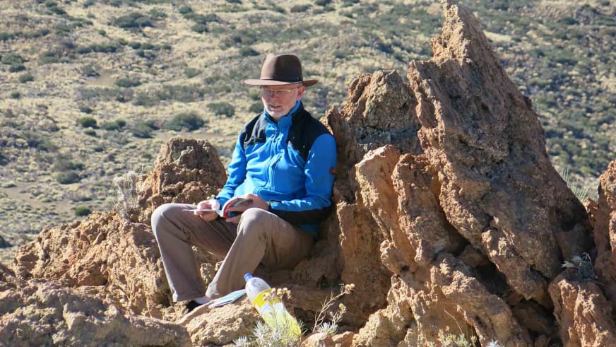 Bergwanderführer Werner Neubauer sitzt lesend auf rotem Sandsteingestein, trägt braunen Hut und blaue Softshelljacke.