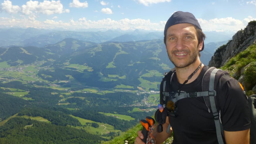 Bergwanderführer Andreas Maier trägt orangefarbene Kletterausrüstung und grauen Rucksack vor Alpenpanorama.