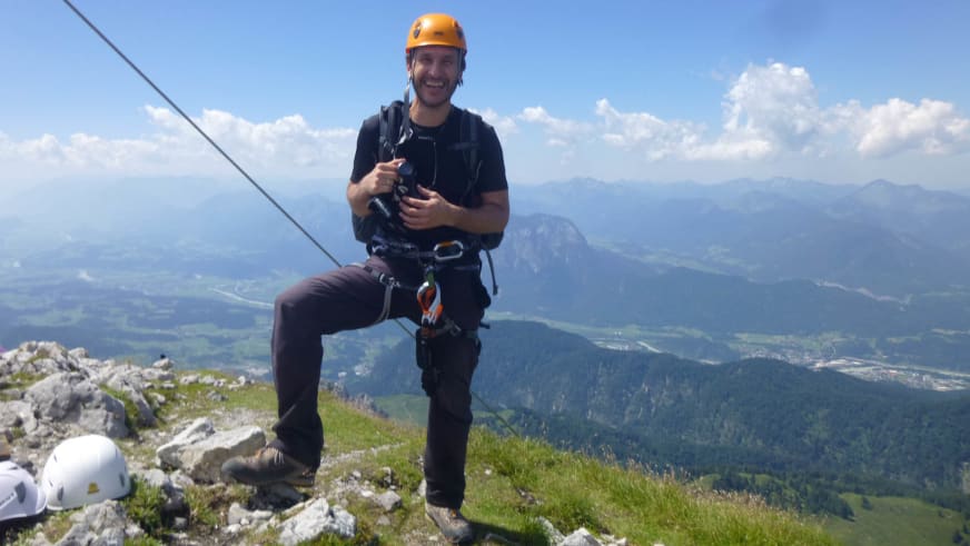 Mit orangem Helm kniet Bergwanderführer Andreas Maier auf einem Berggipfel oberhalb der Wolkendecke.