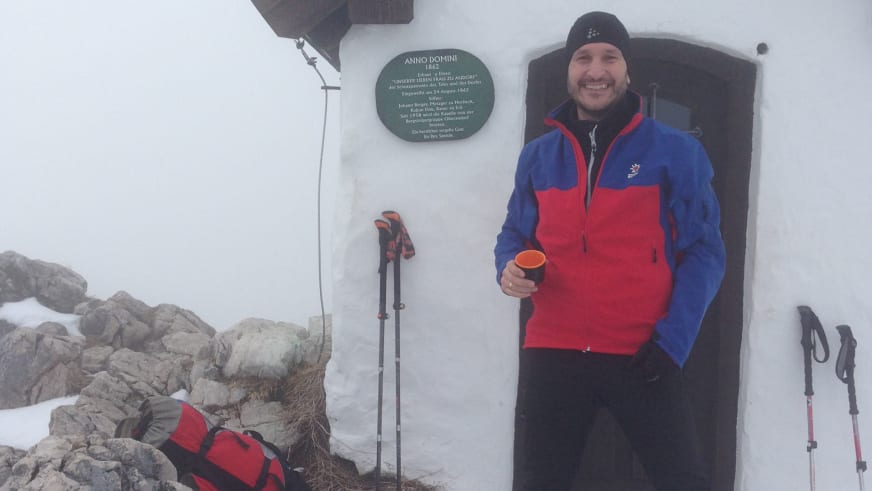 Bergwanderführer Andreas Maier macht Pause vor einer Berghütte mit orangefarbener Tasse in winterlicher Landschaft.