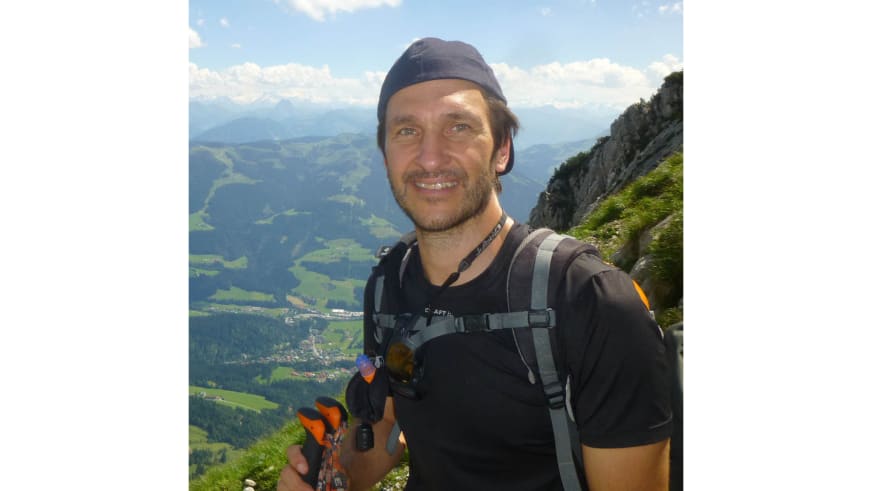 Portrait von Bergwanderführer Andreas Maier mit grauer Kappe und Klettersteig-Ausrüstung vor Alpenlandschaft.