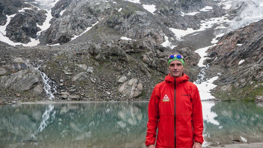 Bergwanderführer Markus Arndt in roter Bergjacke und grüner Mütze vor einem stillen Bergsee mit Felswänden.