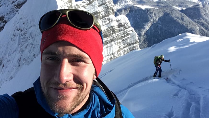 Selfie von Bergführer Julian Conrad mit roter Mütze und Gletscherbrille, im Hintergrund eine zweite Person beim Aufstieg.