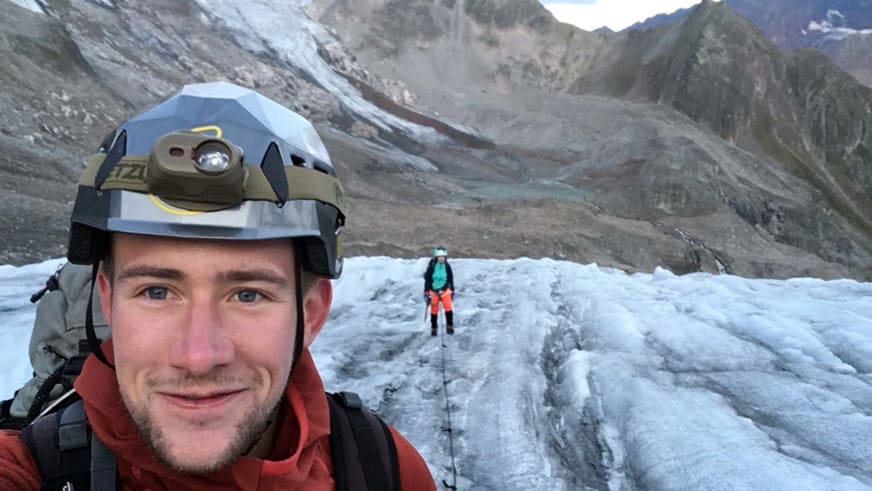 Auf dem Gletscher macht Bergführer Julian Conrad ein Selfie mit Helm und Stirnlampe, eine zweite Person steht mit Skiern dahinter.