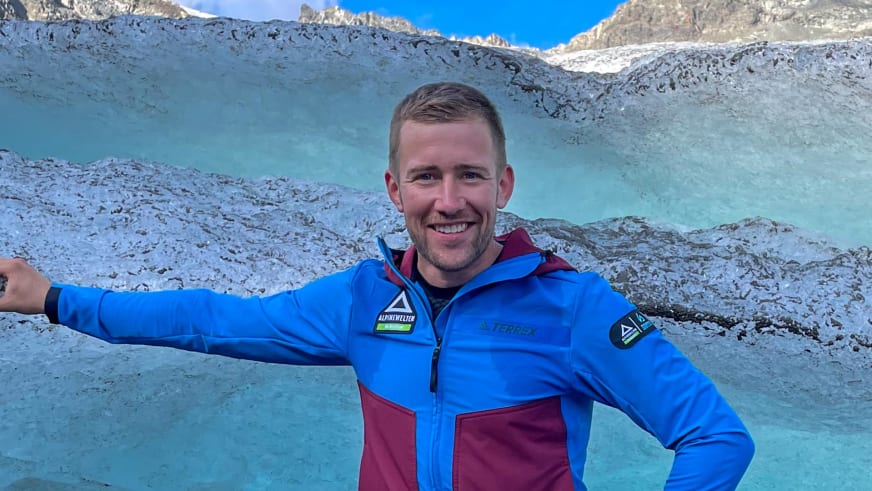 Bergführer Julian Conrad steht lächelnd mit ausgebreiteten Armen vor einem türkisfarbenen Gletschersee in blauer Softshell-Jacke.