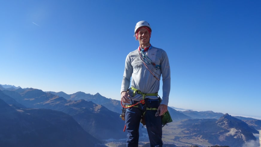 Bergführer Florian Lorenz steht lächelnd mit weißem Helm und grün-gelbem Klettergurt auf dem Watzmann vor einem Bergpanorama.