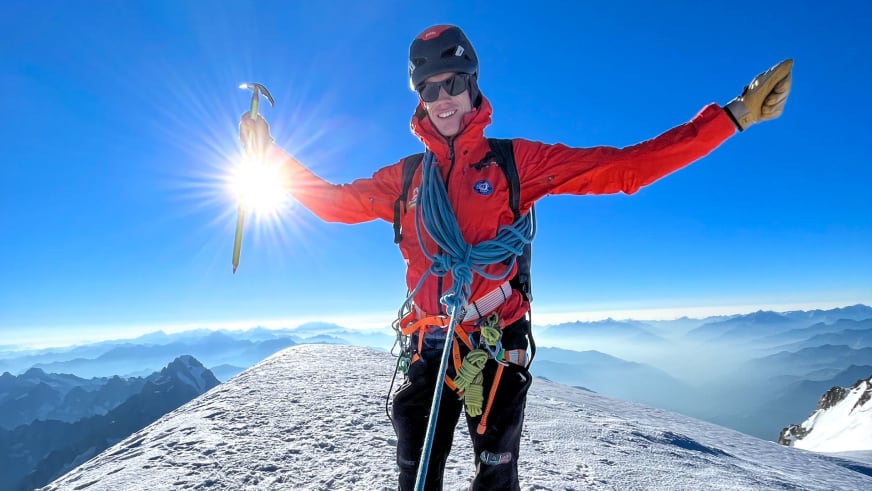 Mit ausgebreiteten Armen steht Bergführer Florian Lorenz in roter Jacke und schwarzem Helm siegreich auf einem verschneiten Gipfel.
