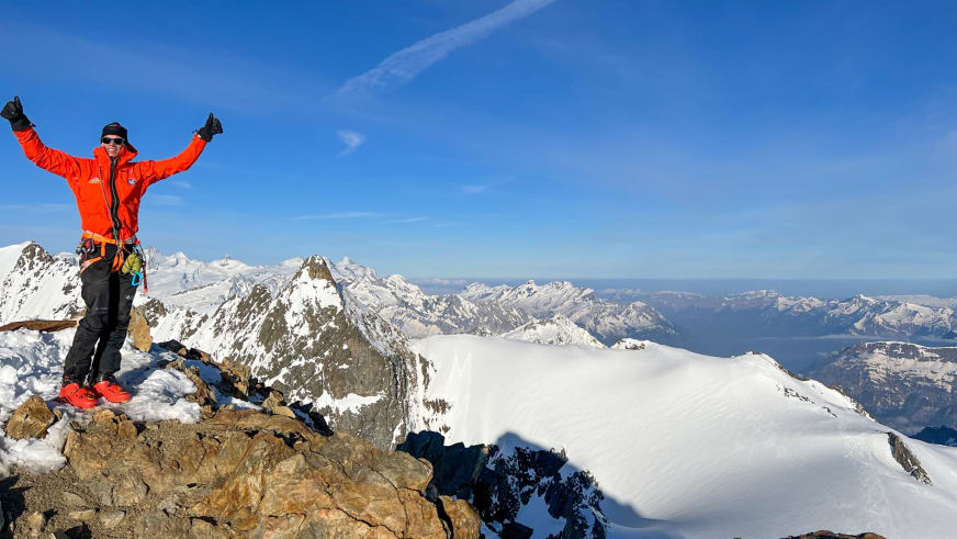 Bergführer Florian Lorenz steht mit ausgestreckten Armen jubelnd auf einem schneebedeckten Felsgipfel in orangeroter Winterjacke.