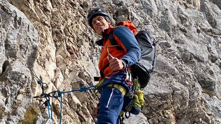 Bergführer Florian Lorenz zeigt Daumen hoch und hält ein blaues Klettersteig-Set an einer hellen Kalksteinwand in orangener Jacke.