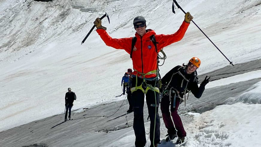 Bergführer Florian Lorenz jubelt mit roter Softshelljacke und Eispickeln auf einem Gletscher, während drei weitere Personen mit alpiner Ausrüstung dahinter stehen.