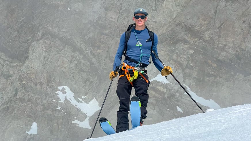 Bergführer Florian Lorenz steht mit Tourenskiern und blauem Alpine Welten Shirt lächelnd vor einer grauen Felswand auf einem Firnfeld.