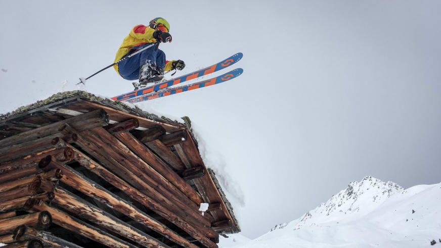 Bergführer Paul Lohse in gelber Jacke springt mit Skiern von einem schneebedeckten Holzdach ab.