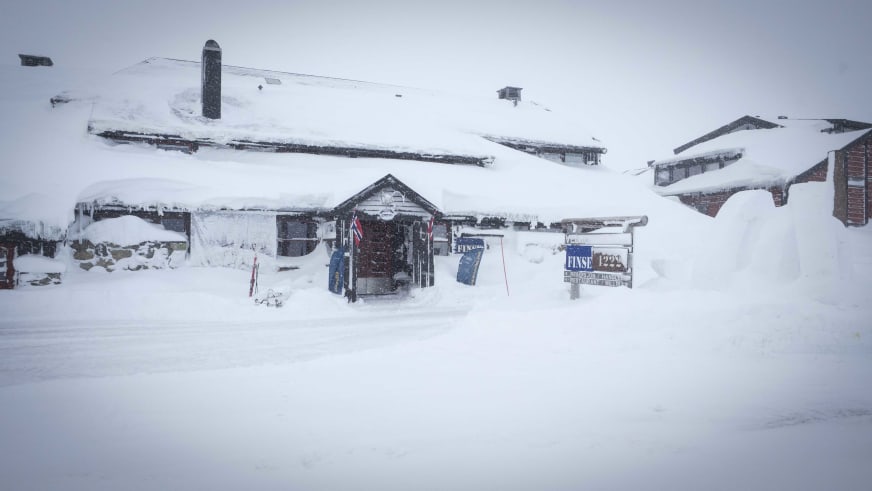 Das Hotel Finse 1222 bei starkem Schneefall mit schneeverdeckten Dächern und dem blauen Hotelschild davor.