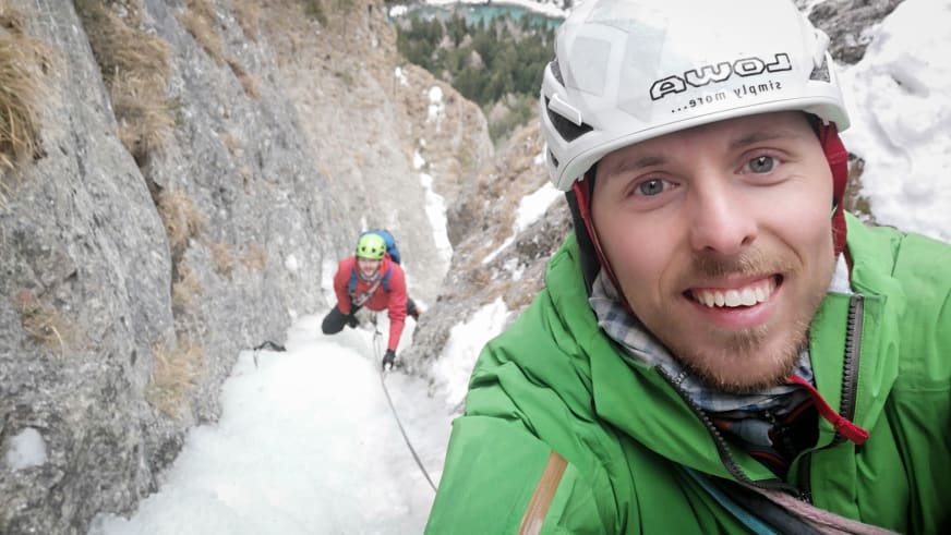 Im Vordergrund lächelt Bergführer Paul Lohse in die Kamera, während eine Person an der schneebedeckten Felswand klettert.