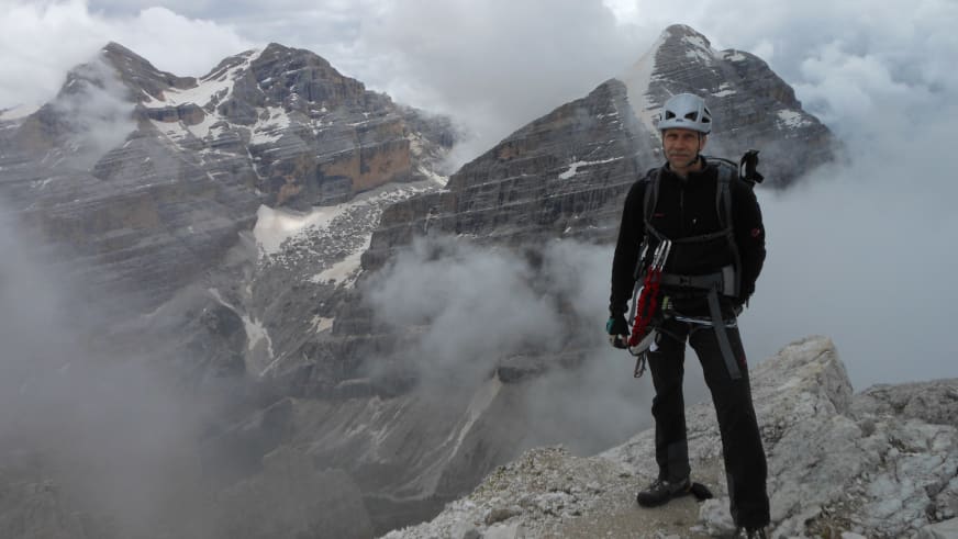 Bergwanderführer Thomas Schill in schwarzer Kleidung und weißem Kletterhelm steht auf felsigem Grat vor verschneiter Berglandschaft.