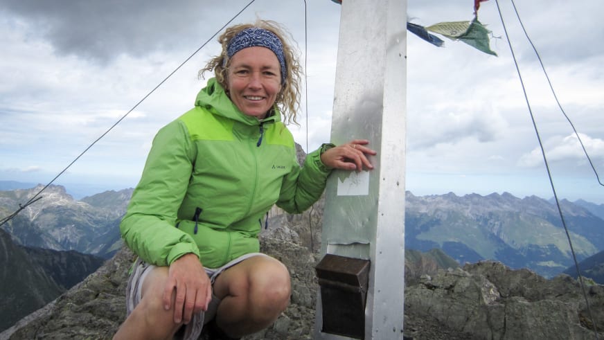Bergwanderführerin Friedlinde Hillmann sitzt am weißen Gipfelkreuz und berührt es mit der Hand vor Bergkulisse.