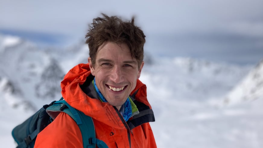 Lächelnder Bergführer Korbinian Schmidtner trägt eine orange-rote Winterjacke vor schneebedeckter Bergkulisse.