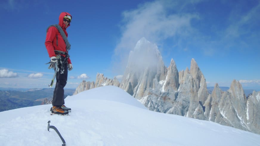 Auf einer Hochtour steht Bergführer Korbinian Schmidtner mit Eispickel auf dem Gipfel vor hellen Kalksteinwänden.